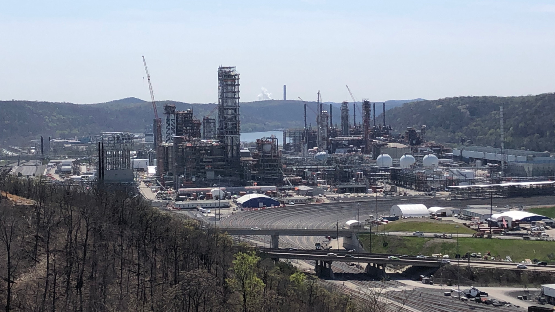 A massive plastic factory sits next to a river on the right and a wooded hill on the left.