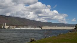 Dam stretching across wide river with wind turbines on hillsides.