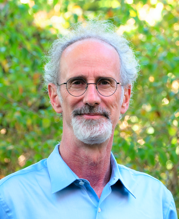 Photo of white man with gray curly hair and black glasses wearing a blue shirt.