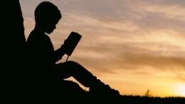A silhouette of a young boy reading a book in the setting sun.