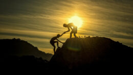 One man helps another up a mountain as the sun sets behind them.