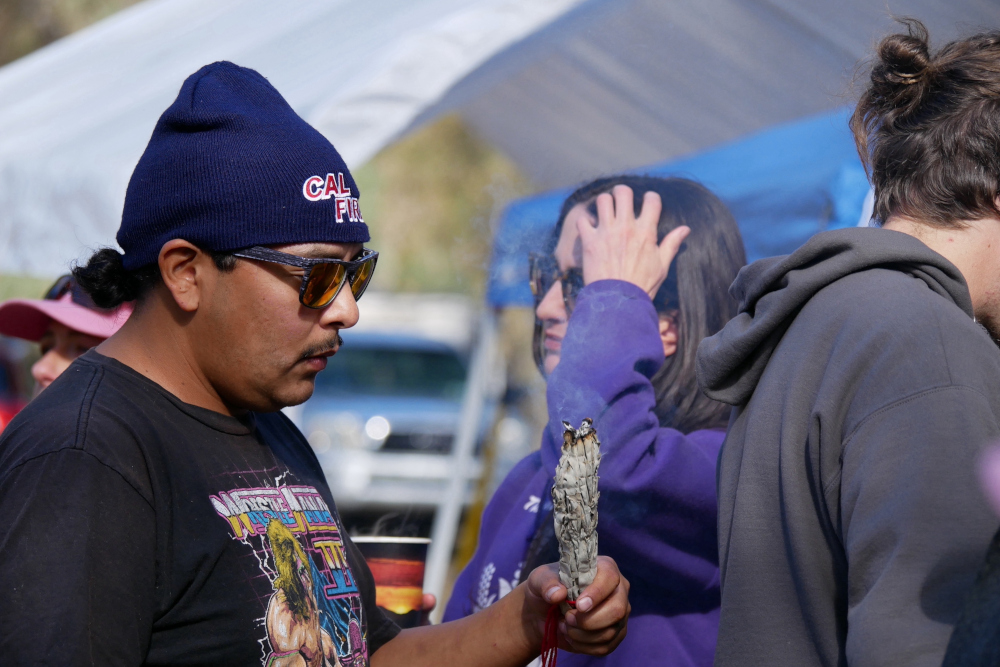 A man in a wrestling tshirt holds burning sage while two other people can be seen in the background
