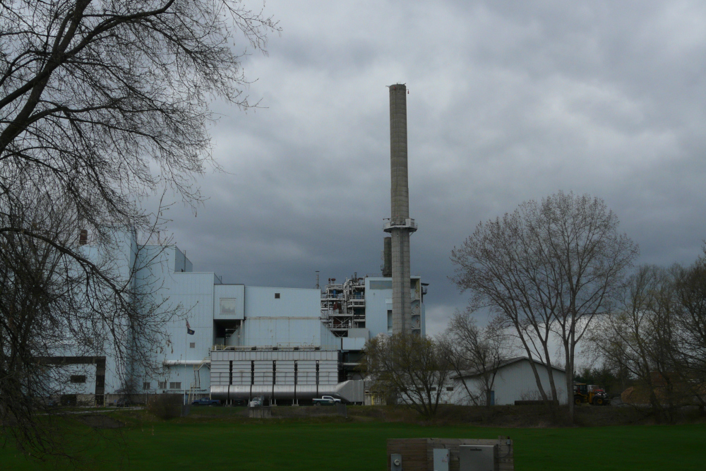 A series of off-white buildings and a tall smokestack against a cloudy sky. A few bare trees on either side.