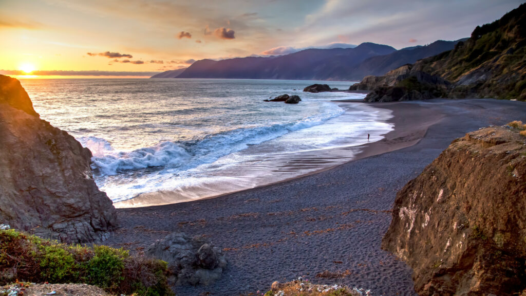 gentle waves fall against a sandy beach, with rocky hills surrounding and the setting sun in the distance