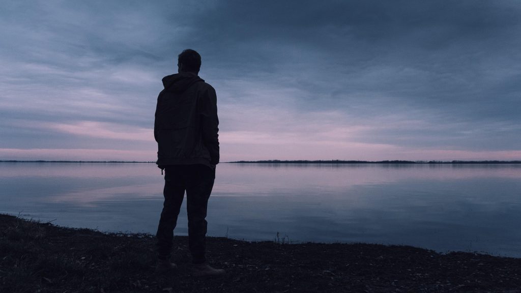 A lone man stares over the water at twilight