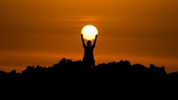 A person silhouetted at sunset, with their arms up as if they are holding the sun