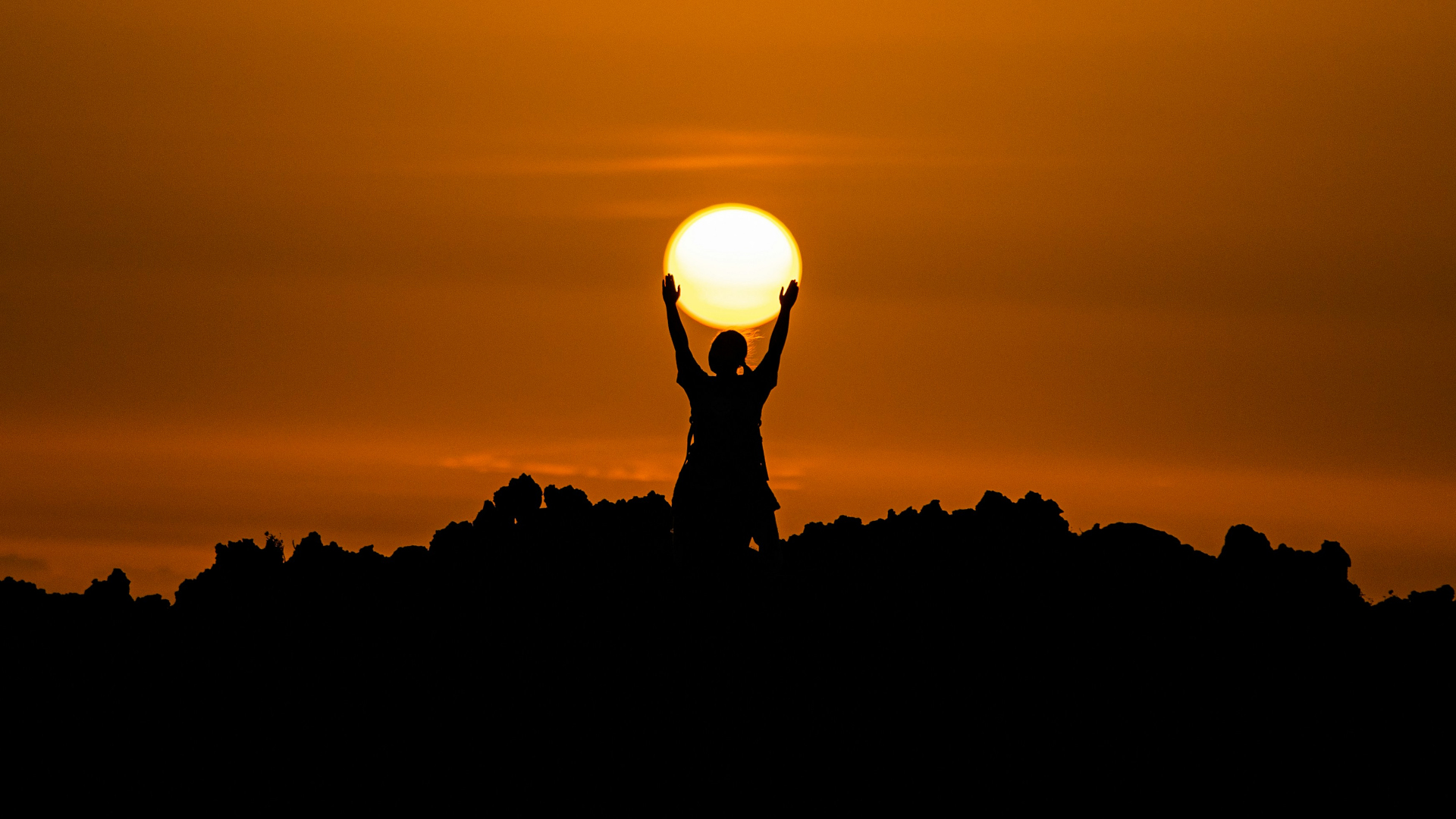 A person silhouetted at sunset, with their arms up as if they are holding the sun