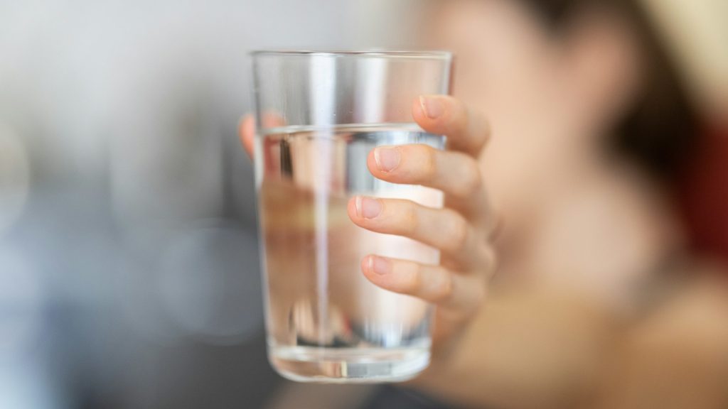 A hand holds a glass of water. The background is out of focus.